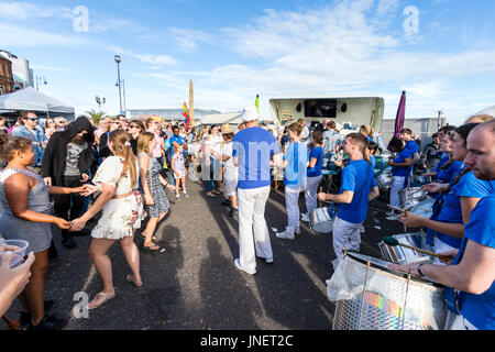 Ritmi della città banda samba eseguendo sul Porto di Ramsgate lungomare di sole splendente, mentre nella parte anteriore dei membri del pubblico su di loro i piedi a ballare. Vista laterale con banda sul diritto pubblico, sulla sinistra. Foto Stock