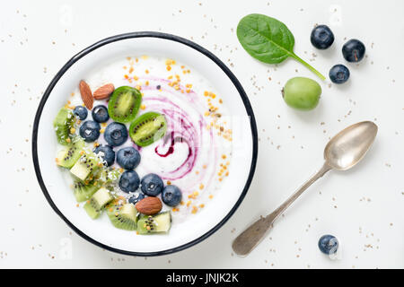 Yogurt con mirtilli e kiwi, spinaci, mandorle, scaglie di cocco e polline di api in una ciotola sul tavolo bianco. Vista superiore Foto Stock