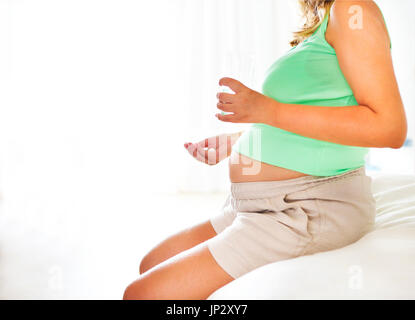 Giovane donna incinta con la medicina e bicchiere di acqua seduta sul letto in camera Foto Stock