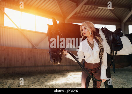 Bellissima ed elegante ragazza bionda sta in piedi vicino al suo cavallo nero medicazione di concorrenza uniforme camicetta bianca maglietta e pantaloni marrone. Piscina portraite in Foto Stock