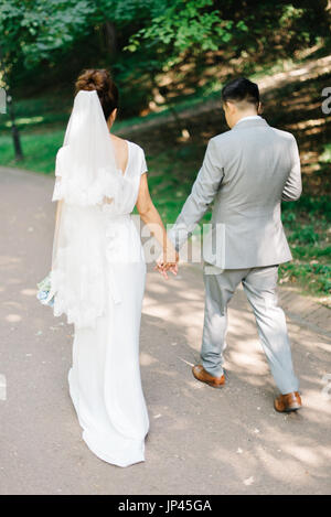 Lo sposo in abito grigio detiene sposa la mano mentre si cammina in estate Foto Stock