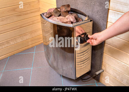 Sauna elettrico riscaldatore stufa con pietre poste sulla sommità di esso, la mano gira il regolatore sul pannello di comando Impostazione del tempo e della temperatura. Foto Stock