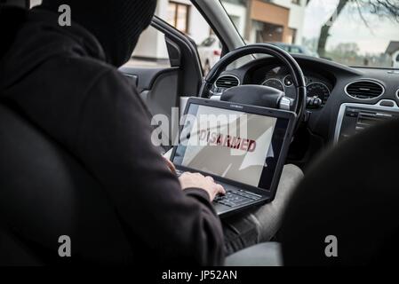 L'uomo vestito di nero e con un passamontagna sul suo capo disarmante protezioni auto con computer portatile. Ladro di auto, il furto di auto concept Foto Stock