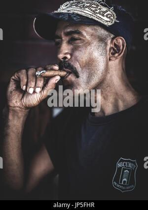 Un uomo che indossa un cappello da baseball di fumare un sigaro. Foto Stock