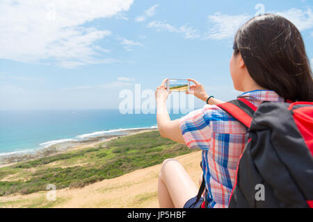 Vista posteriore foto della femmina azienda backpacker telefono cellulare utilizza la tecnologia fotocamera per scattare una bella vista del mare e delle colline seduti sulla montagna relax. Foto Stock