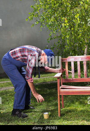 Senior uomo in tuta la verniciatura vecchia panca in giardino dopo la sabbiatura. Riparazione di mobili vecchi Foto Stock