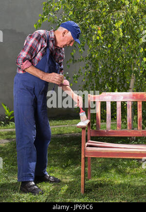 Senior uomo in tuta la verniciatura vecchia panca in giardino dopo la sabbiatura. Riparazione di mobili vecchi Foto Stock