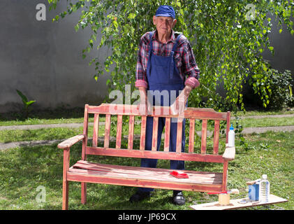 Senior uomo in tuta la verniciatura vecchia panca in giardino dopo la sabbiatura. Riparazione di mobili vecchi Foto Stock