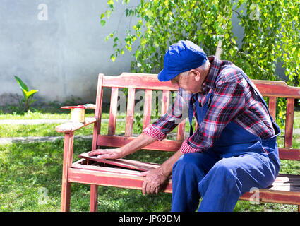 Senior uomo in tuta la verniciatura vecchia panca in giardino dopo la sabbiatura. Riparazione di mobili vecchi Foto Stock