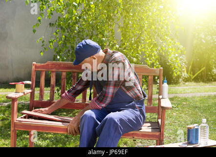 Senior uomo in tuta la verniciatura vecchia panca in giardino dopo la sabbiatura. Riparazione di mobili vecchi Foto Stock