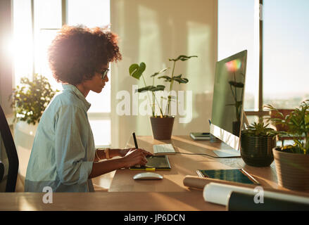 Vista laterale della femmina di graphic designer che lavora con disegno digitale tavoletta e penna su un computer in ufficio. Donna Africana a lavorare alla sua scrivania. Foto Stock