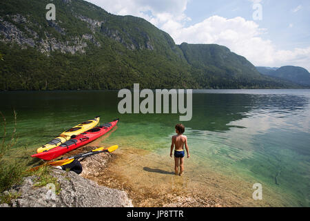 Giovane ragazzo in piedi da due kayak nelle calme acque chiare del lago alpino di Bohinj, Slovenia. Foto Stock