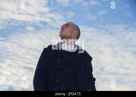 Bald giovane con occhi marroni vestito in moderne ed eleganti vestiti sul cielo azzurro sfondo con nuvole. Foto Stock