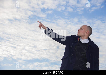 Bald giovane con occhi marroni vestito in moderne ed eleganti vestiti sul cielo azzurro sfondo con nuvole. Foto Stock