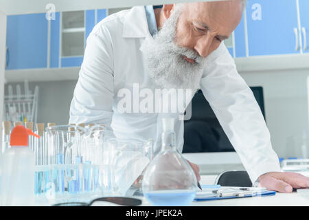 Concentrato barbuto senior scientist in camice bianco in scrittura di appunti su tanle con palloni in laboratorio chimico Foto Stock