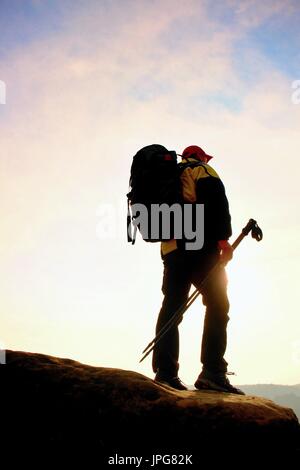 Silhouette di turista con cappuccio rosso e pali in mano. Escursionista con zaino grande stand su roccia. Molla di sole lo spuntar del giorno nelle montagne rocciose. Foto Stock