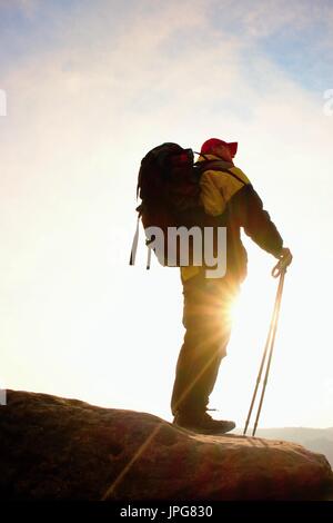 Silhouette di turista con cappuccio rosso e pali in mano. Escursionista con zaino grande stand su roccia. Molla di sole lo spuntar del giorno nelle montagne rocciose. Foto Stock