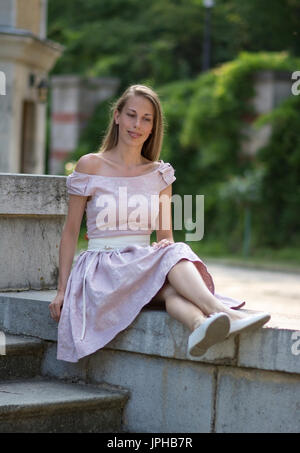 Ritratto di bella ragazza bianco in abito rosa seduta sulla vecchia scala di pietra. Foto Stock