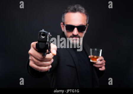 I Gangster uomo che ha di mira con la pistola alla fotocamera e bere whisky raffinati Foto Stock