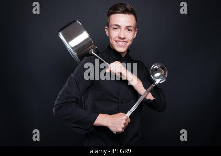 Allegro chef con utensili da cucina sempre pronto a preparare un buon pasto su sfondo scuro Foto Stock