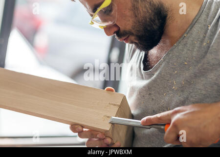 Un dai capelli scuri con barba protettivi e occhiali verdi il trattamento di un prodotto di legno con uno scalpello in officina, close-up, vista laterale Foto Stock