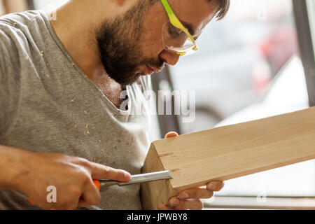 Un dai capelli scuri con barba protettivi e occhiali verdi il trattamento di un prodotto di legno con uno scalpello in officina, close-up, vista laterale Foto Stock