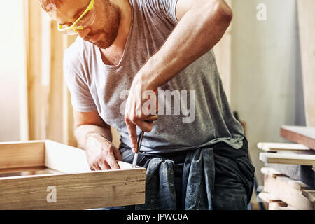 Un dai capelli scuri con barba protettivi e occhiali verdi il trattamento di un prodotto di legno con uno scalpello in officina Foto Stock