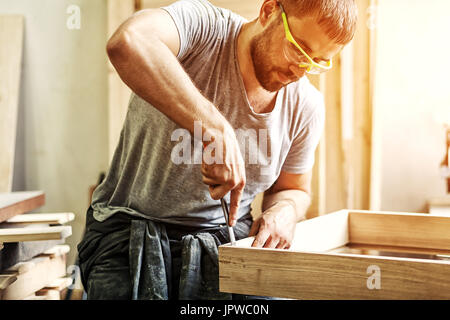 Un dai capelli scuri con barba protettivi e occhiali verdi il trattamento di un prodotto di legno con uno scalpello in officina Foto Stock