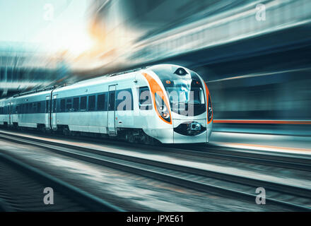 Treno ad alta velocità in movimento con la stazione ferroviaria al tramonto in Europa. Moderno treno intercity sulla linea ferroviaria della piattaforma con moto effetto di sfocatura Foto Stock