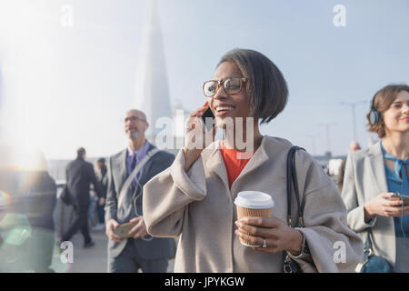 Sorridente imprenditrice a bere caffè e parlando al cellulare sul soleggiato urbano occupato un ponte pedonale Foto Stock