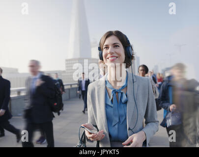 Sorridente, fiduciosi imprenditrice ascoltando musica con smart phone e cuffie su urban ponte pedonale Foto Stock