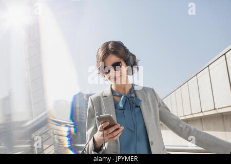 Imprenditrice sorridente con occhiali da sole l'ascolto di musica con smart phone e cuffie Foto Stock