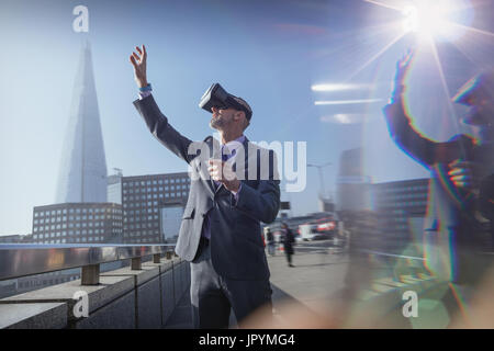 Imprenditore utilizzando la realtà virtuale occhiali simulatore, raggiungendo per sky sul soleggiato ponte urbano, London, Regno Unito Foto Stock