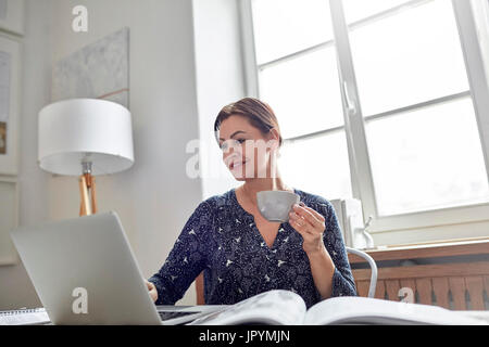 Sorridente imprenditrice a bere caffè e lavorare al computer portatile Foto Stock