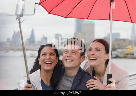 Sorridente amico turisti con ombrello tenendo selfie con selfie stick, London, Regno Unito Foto Stock