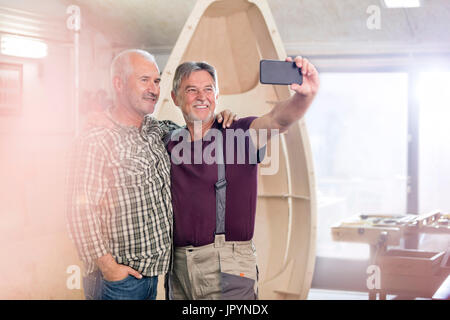 Orgoglioso e sorridente falegnami maschio con la fotocamera del telefono tenendo selfie avanti troppo barca di legno in officina Foto Stock