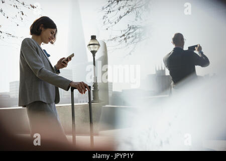 Imprenditrice con valigia tramite telefono cellulare nel soleggiato parco urbano, London, Regno Unito Foto Stock