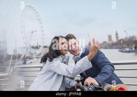 Coppia sorridente turisti tenendo selfie con la fotocamera del telefono sul ponte in prossimità di Millennium Wheel, London, Regno Unito Foto Stock