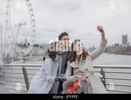 Sorridono felici amici prendendo selfie con selfie stick sul ponte in prossimità di Millennium Wheel, London, Regno Unito Foto Stock