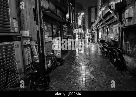 TOKYO, Giappone - 8 Aprile 2017 - In bianco e nero di un uomo con un ombrello a piedi verso il basso un solitario vicolo su una notte piovosa nella città di Tokyo Foto Stock