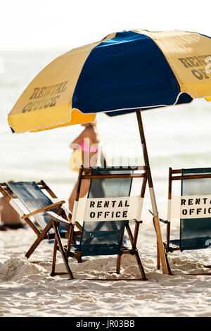 Sdraio e ombrellone sulla spiaggia. Una donna in bikini che cammina sullo sfondo. Foto Stock