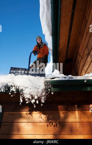 Una giovane donna rimuove la neve dal tetto della sua casa; Omero, Alaska, Stati Uniti d'America Foto Stock