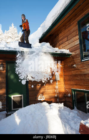 Una giovane donna rimuove la neve dal tetto della sua casa; Omero, Alaska, Stati Uniti d'America Foto Stock