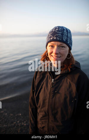 Ritratto di una donna con i capelli rossi che indossa un cappello di maglia e permanente al bordo dell'acqua; Alaska, Stati Uniti d'America Foto Stock