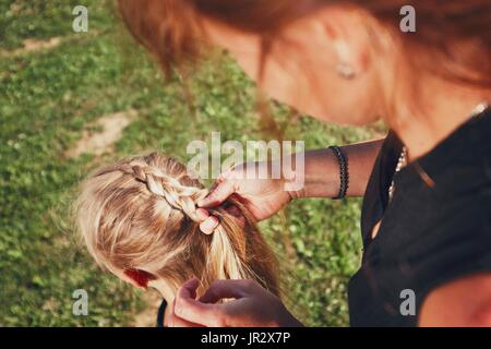 Giovane donna rendendo trecce di capelli della bambina sul giardino in campagna. Foto Stock