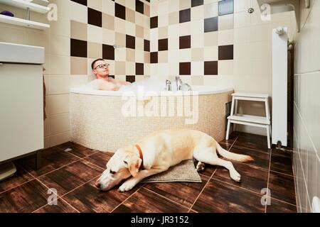 Vivere con il cane. Giovane uomo relax nel bagno con il suo giallo labrador retriever. Foto Stock