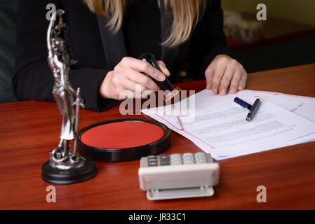 Primo piano sulla mano di notaio timbro sul documento. Accanto al notaio statuetta della dea della giustizia - Themis Foto Stock