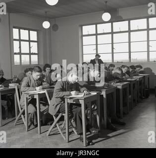 Anni '50, Inghilterra, giovani studenti adulti seduti a piccole scrivanie individuali in legno in un corso di un college tecnico. Scrivono su carta con penne, mentre un'insegnante femminile controlla il lavoro, Inghilterra, Regno Unito. I giovani maschi indossano giubbotto e cravatte, un standard formale di abbigliamento comune a questa era post-bellica. Foto Stock