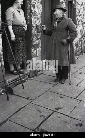 1940s, storico, uomo anziano con indossando un soprabito e cappello e di tenere su un bastone da passeggio, chat per una signora all'ingresso al suo cottage circa il suo acquisto di alcuni dei suoi cardidi. Foto Stock