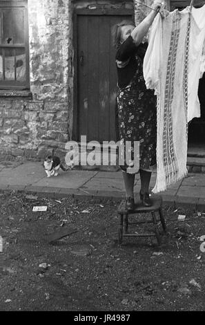 1940s, Merthyr Tydfil, South Wales, signora in grembiule in piedi su uno sgabello in legno appesi panni bagnati ad asciugare in strada al di fuori del suo cottage in pietra. Foto Stock